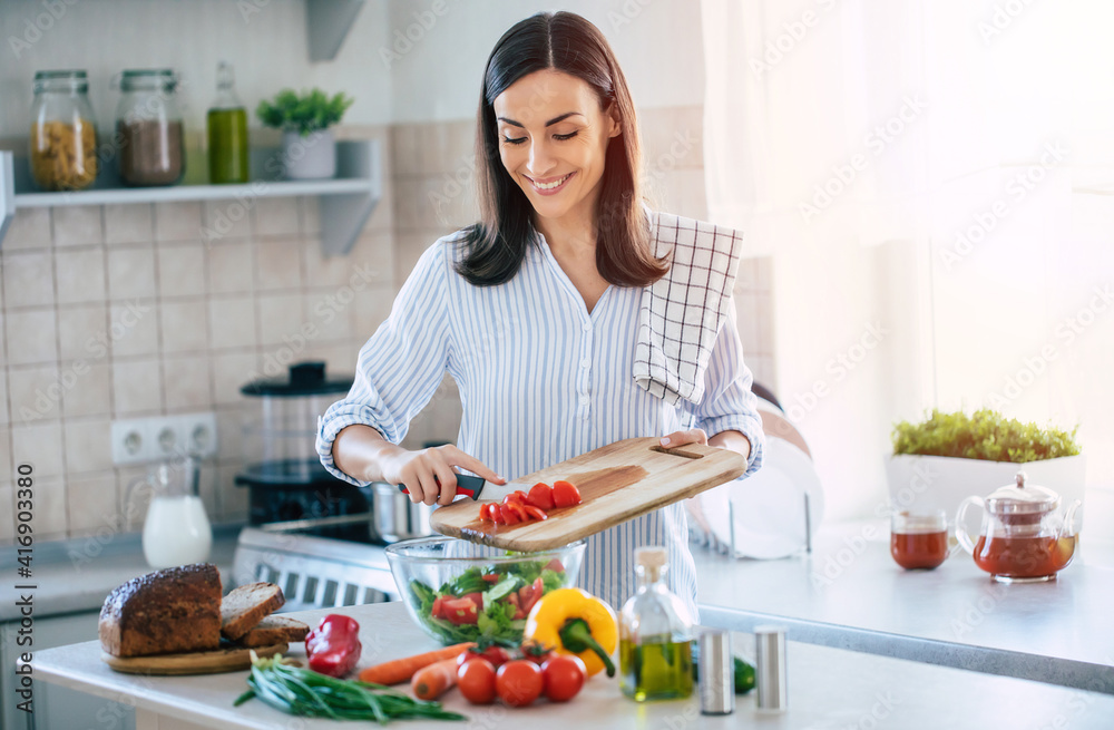 Happy smiling cute woman is preparing a fresh healthy vegan salad with many vegetables in the kitchen at home and trying a new recipe