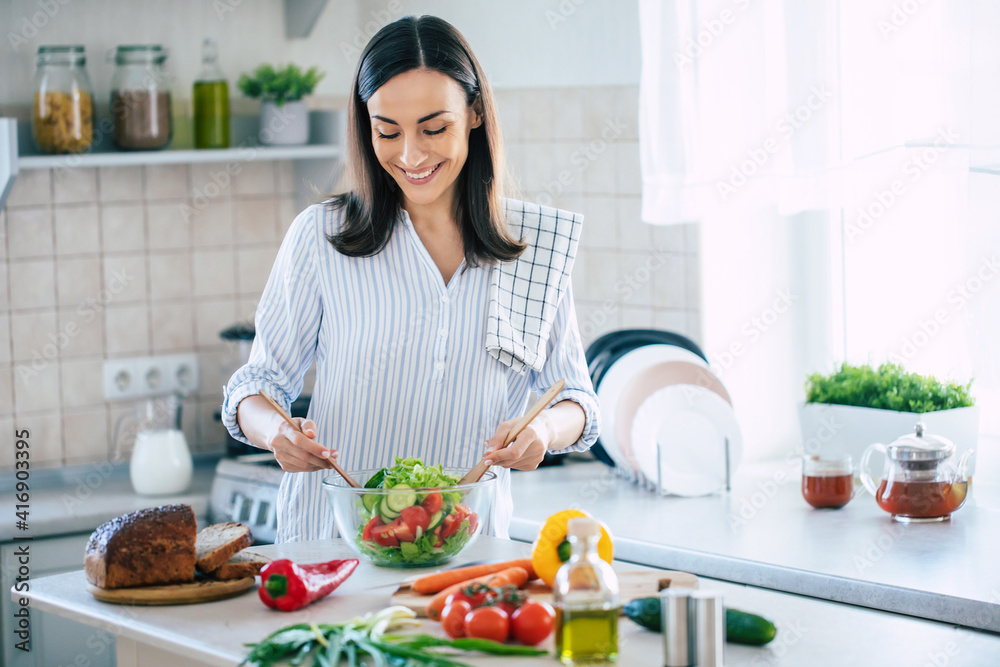 Happy smiling cute woman is preparing a fresh healthy vegan salad with many vegetables in the kitchen at home and trying a new recipe