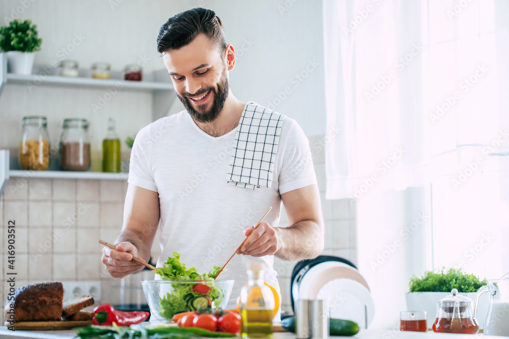 Handsome happy bearded man is preparing wonderful fresh vegan salad in the kitchen at home Handsome happy bearded man is preparing wonderful fresh vegan salad in the kitchen at home