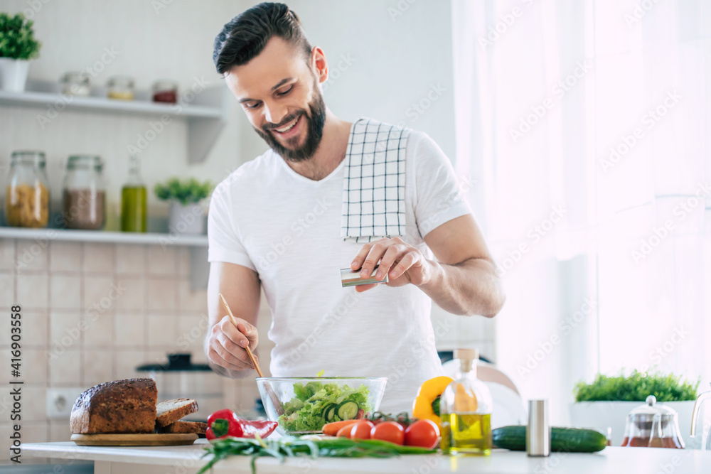 Handsome happy bearded man is preparing wonderful fresh vegan salad in the kitchen at home Handsome happy bearded man is preparing wonderful fresh vegan salad in the kitchen at home