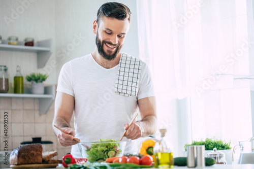 Handsome happy bearded man is preparing wonderful fresh vegan salad in the kitchen at home