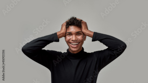 Joyful young guy smiling at camera and holding his head in surprise, posing isolated over gray background