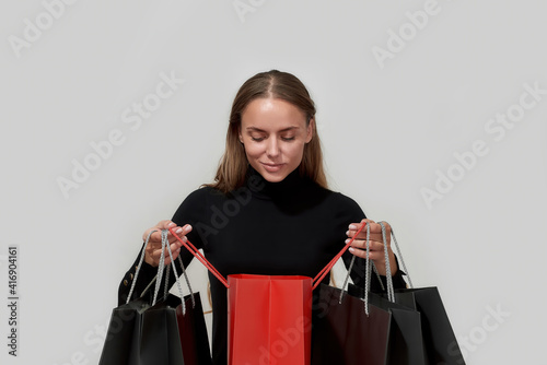 Portrait of curious young caucasian woman dressed in black looking inside one of many shopping bags while standing isolated over light gray background