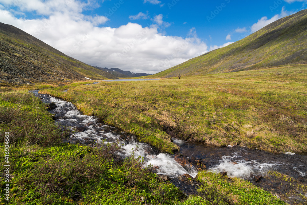 Beautiful summer arctic landscape. View of a small stream in the tundra, flowing from the lakes in the valley between the hills. In the distance, a small figure of a fisherman with a fishing rod.
