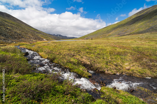 Beautiful summer arctic landscape. View of a small stream in the tundra, flowing from the lakes in the valley between the hills. In the distance, a small figure of a fisherman with a fishing rod.