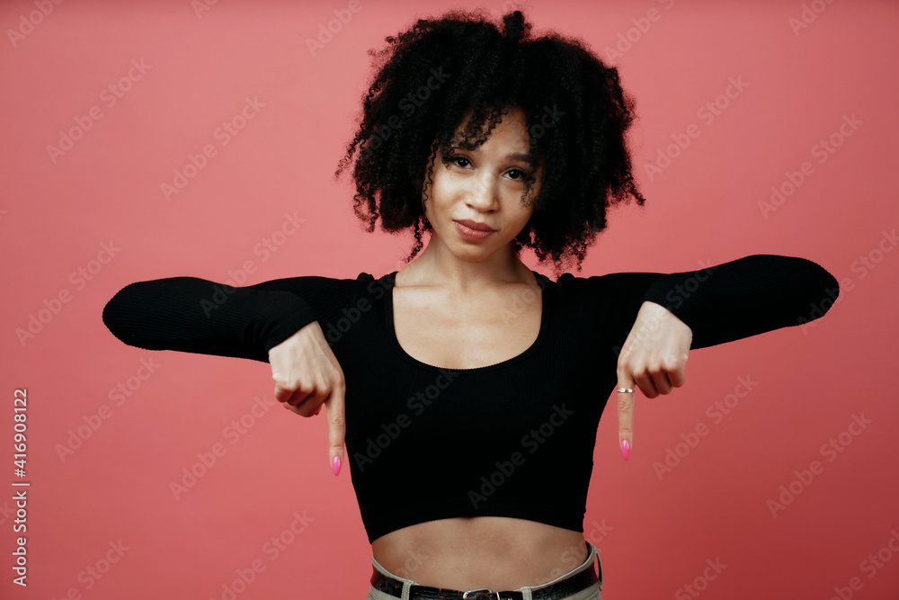 makes a downward gesture with her hands, a young Afro-ethnic woman. A model in a photo studio poses on a scarlet background.