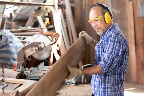 senior asian man carpenter wearing glasses and headphone, holding looking and aiming wooden plank in carpentry workshop