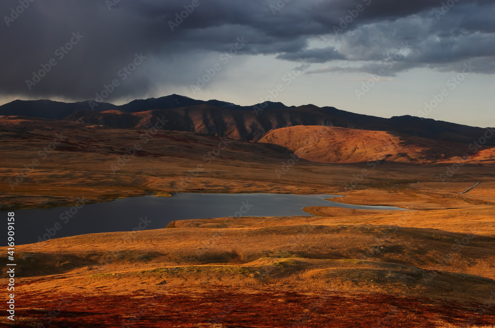 Colorful sunset above highland steppe shore of lake with dry yellow grass on the background of rocky mountains under storm sky