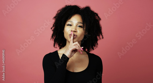 shows a finger gesture of quiet silence to the camera and smiles, a young woman of Afro appearance. photo in a photo studio on a red background.