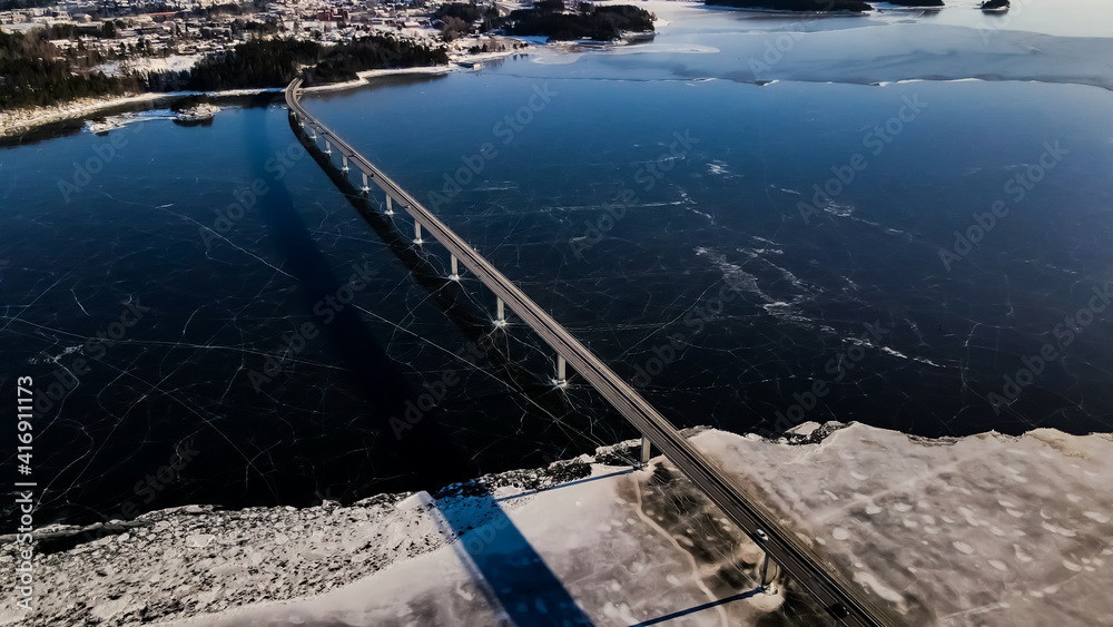 Traffic driving on a bridge over a bay covered in clear thick ice with cracked pattern. Aerial photo.