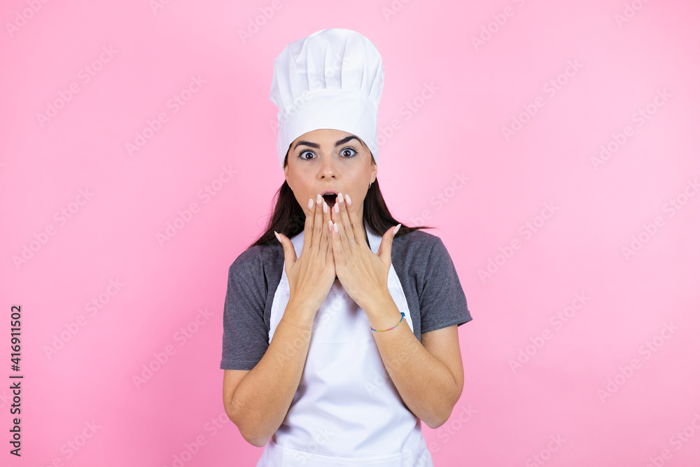 Young hispanic woman wearing baker uniform over pink background shocked covering mouth with hands for mistake. secret concept.