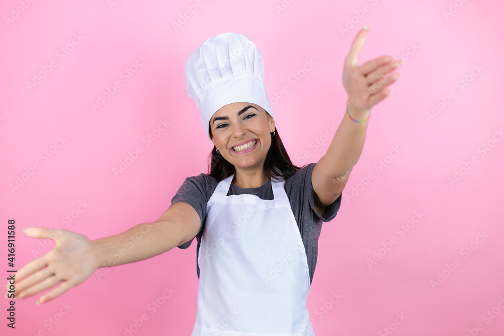 Young hispanic woman wearing baker uniform over pink background looking at the camera smiling with open arms for hug Young hispanic woman wearing baker uniform over pink background looking at the camera smiling with open arms for hug