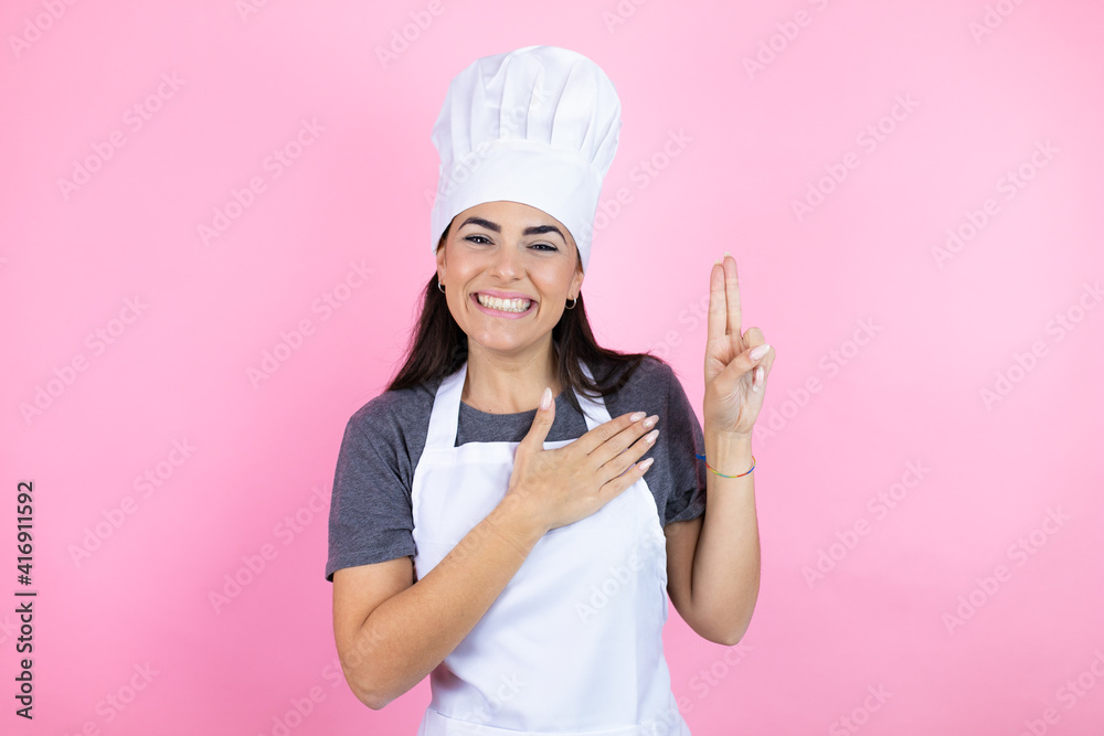 Young hispanic woman wearing baker uniform over pink background smiling swearing with hand on chest and fingers up, making a loyalty promise oath Young hispanic woman wearing baker uniform over pink background smiling swearing with hand on chest and fingers up, making a loyalty promise oath