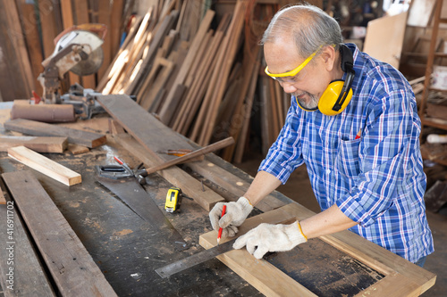 senior asian man carpenter wearing glasses and headphone, measuring wood with ruler in workshop