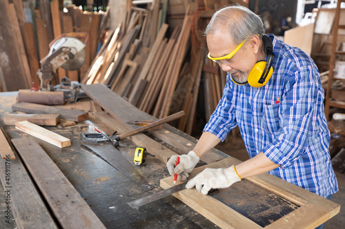 senior asian man carpenter wearing glasses and headphone, measuring wood with ruler in workshop © offsuperphoto