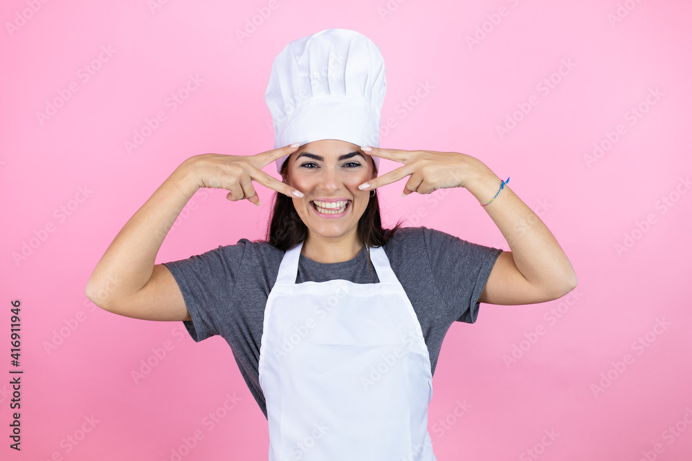 Young hispanic woman wearing baker uniform over pink background Doing peace symbol with fingers over face, smiling cheerful showing victory Young hispanic woman wearing baker uniform over pink background Doing peace symbol with fingers over face, smiling cheerful showing victory