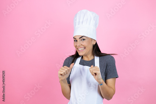 Billede på lærred Young hispanic woman wearing baker uniform over pink background smiling proudly and confidently with her arms on her hip akimbo, happy and confident of success, giving a successful look