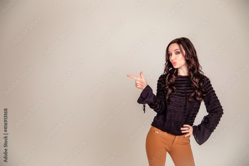 beautiful slender young girl with black long hair on an isolated beige and gray background. woman in brown trousers and black blouse shows with forefinger hand beautiful slender young girl with black long hair on an isolated beige and gray background. woman in brown trousers and black blouse shows with forefinger hand
