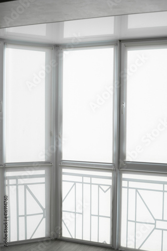 White fabric roller blinds on the plastic window on a balcony in the living room with a reflection in a stretch ceiling.