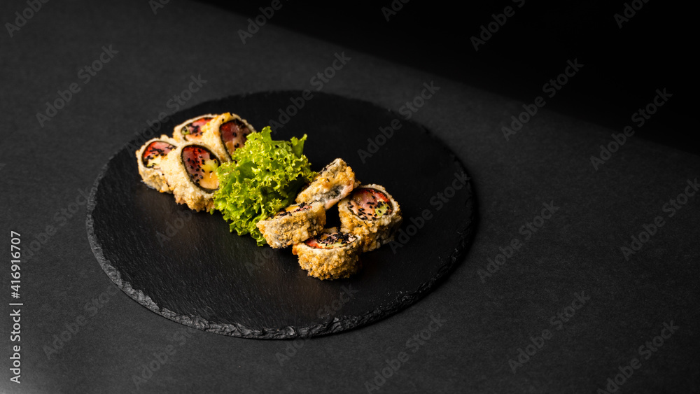 Custom sushi roll in tempura with nori, fresh salmon, tuna, avocado, masago caviar, drizzled with pineapple sauce with salad pouring as decoration on a black plate on a black table and background. Custom sushi roll in tempura with nori, fresh salmon, tuna, avocado, masago caviar, drizzled with pineapple sauce with salad pouring as decoration on a black plate on a black table and background.