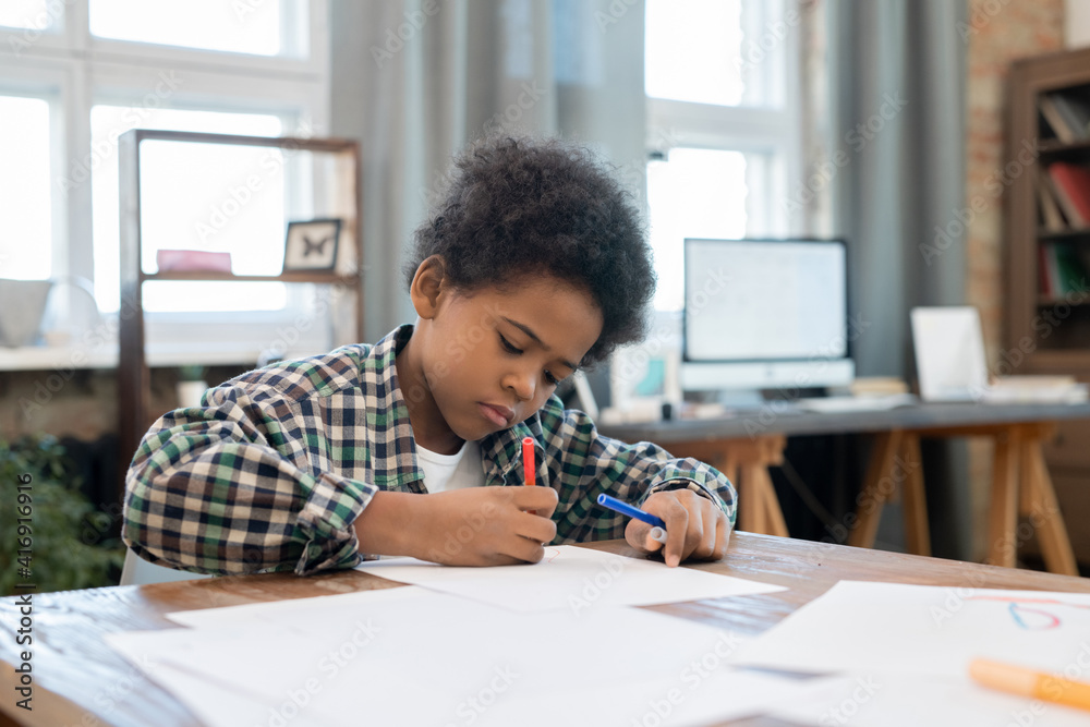 Diligent schoolboy in casualwear drawing with highlighters on paper by table Diligent schoolboy in casualwear drawing with highlighters on paper by table