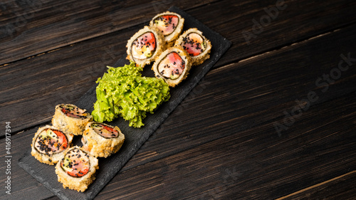 Custom sushi roll in tempura with nori, fresh salmon, tuna, avocado, masago caviar, drizzled with pineapple sauce with salad pouring as decoration on a black plate on a wooden table and background.