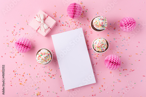 Empty white blank, gift box, paper decorations and cupcakes on pink sprinkles background.