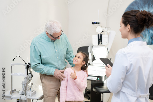 Senior man with his happy granddaughter standing in front of ophthalmologist
