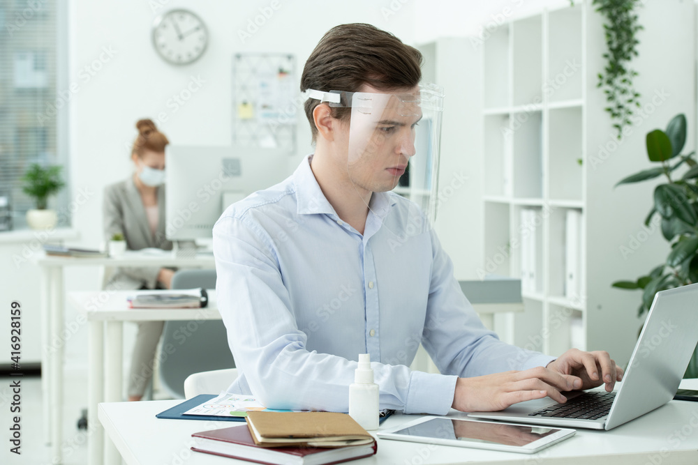 Young serious businessman in protective screen on head looking at laptop display Young serious businessman in protective screen on head looking at laptop display