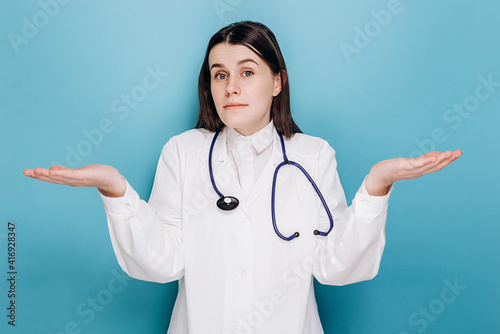 Fotografie Portrait of indecisive woman doctor in white coat, shrugging and looking troubled answer, dont know, isolated on blue wall