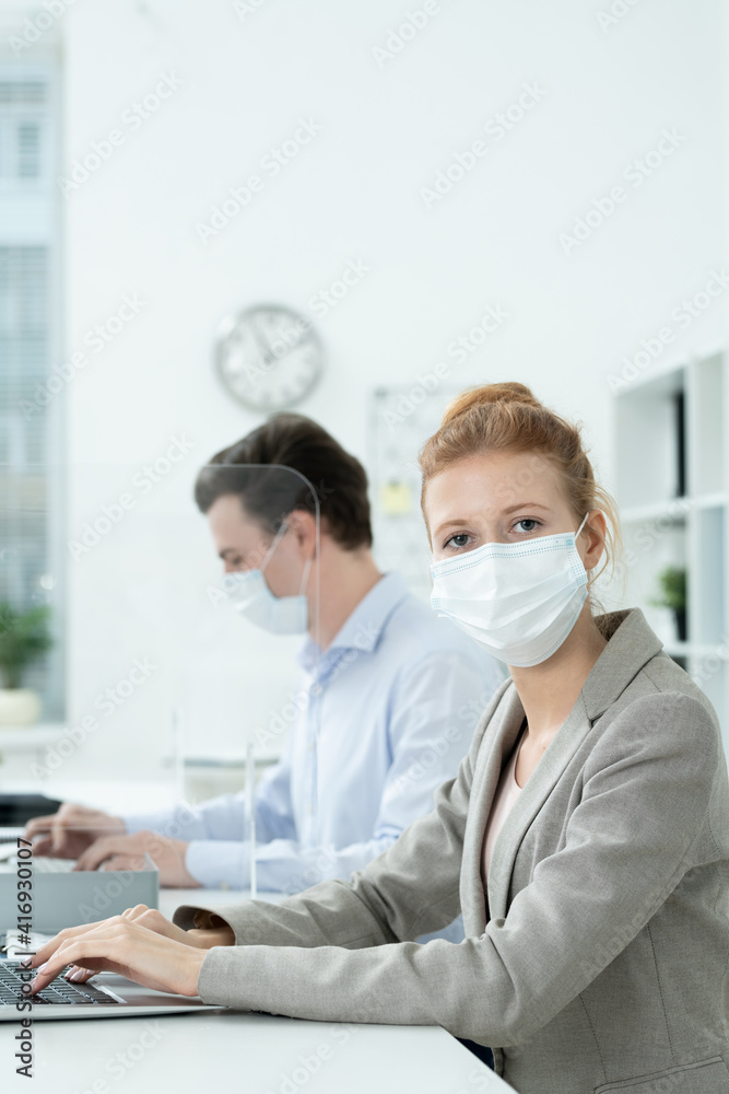 Young female office manager in protective mask and elegant suit using laptop