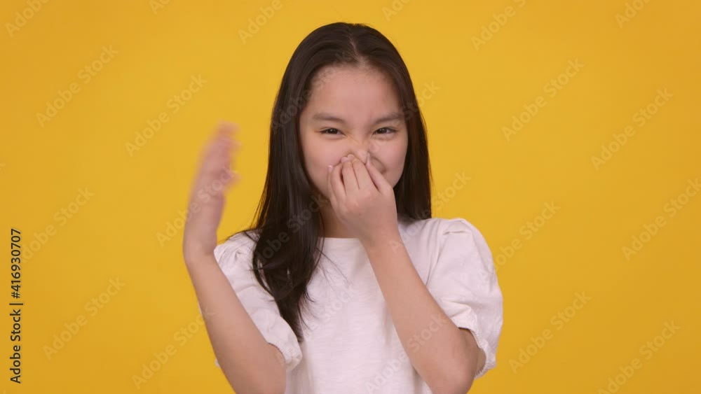 Studio portrait of young frowning girl closing her nose and waving hand, feeling terrible scent, orange background Studio portrait of young frowning girl closing her nose and waving hand, feeling terrible scent, orange background