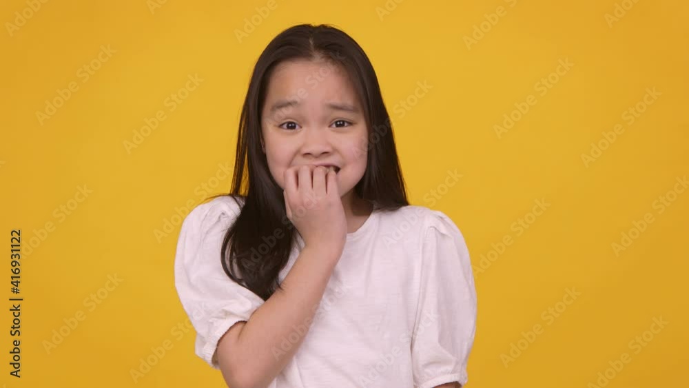 Little asian girl feeling panic, biting her nails nervously, orange studio background, slow motion Little asian girl feeling panic, biting her nails nervously, orange studio background, slow motion