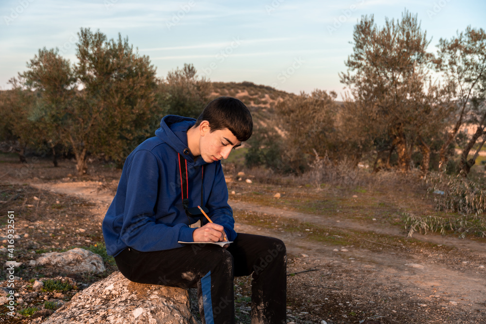 Fototapeta premium Young boy writing in little notebook while sitting in the countryside.