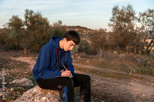 Young boy writing in little notebook while sitting in the countryside. © Manuel