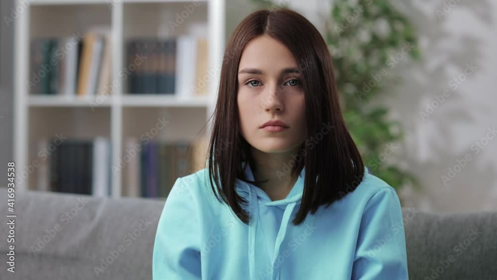 Portrait of young woman sitting on couch with sad facial expression. Unhappy female with brown hair wearing casual blue hoodie. Bad day concept. Portrait of young woman sitting on couch with sad facial expression. Unhappy female with brown hair wearing casual blue hoodie. Bad day concept.
