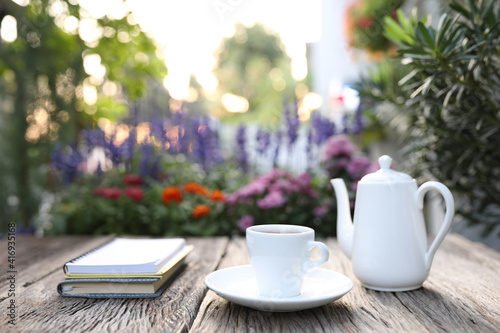 White tea cup and kettle with notebook on wooden table and flower field background
