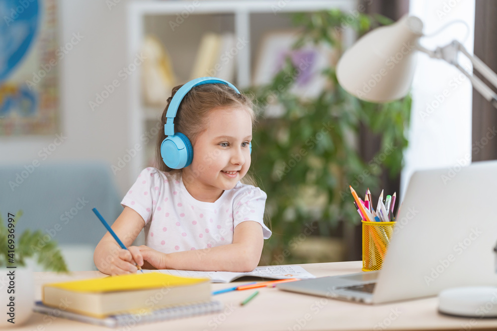 smart smiling little girl wearing headphones is learning online, using laptop remote learning. child listens to teacher and writes down task in notebook. interactive E-Education Distance Home learning smart smiling little girl wearing headphones is learning online, using laptop remote learning. child listens to teacher and writes down task in notebook. interactive E-Education Distance Home learning