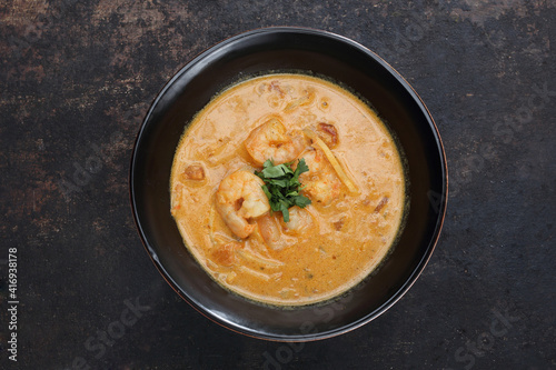 An oriental soup with tomatoes, coconut milk, shrimps, bamboo shoots, on a black stone background, top view.