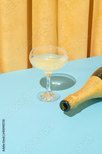 Minimal retro vintage scene with champagne glass and bottle on pastel blue table against yellow velvet curtain. Bright light, retro party.