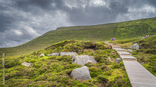 People hiking on wooden boardwalk, between boulders and heathers, leading to Cuilcagh Mountain. Storm, dramatic sky in background, Northern Ireland photo