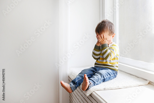 Portrait of adorable little boy sitting on the windowsill and crying. Upset child covering his face at home. Barefoot kid hiding behind palms of his hands. Close up, copy space, background.