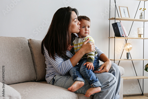 Portrait of adorable little boy sitting on his mom's laps and crying. Mother comforting upset toddler with a kiss on a cheek. Parental love concept. Close up, copy space, background.