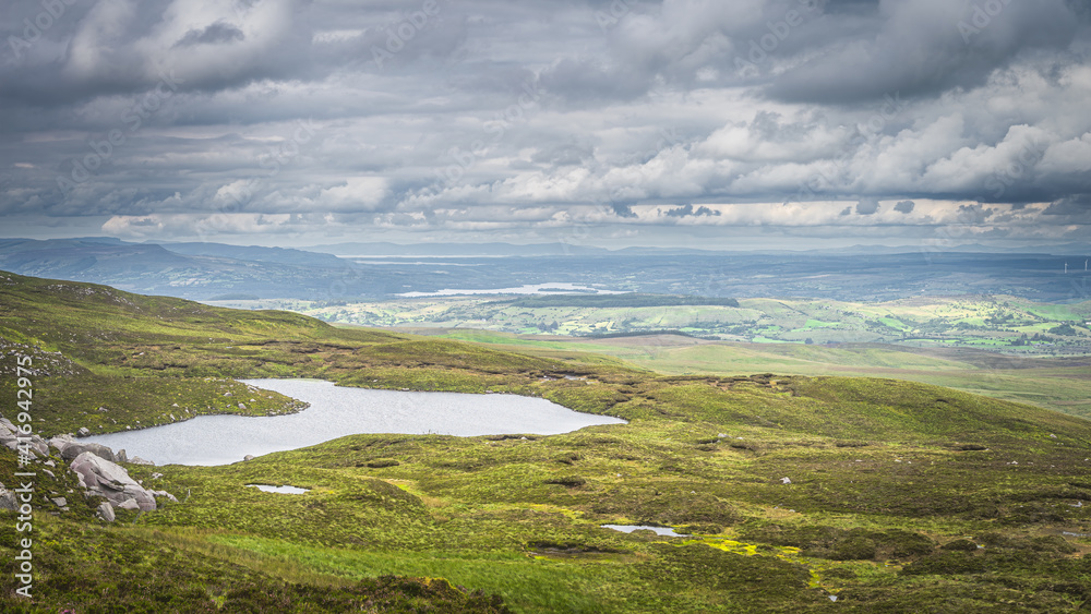 Green fields or rolling hills with small lake at footstep of Cuilcagh Mountain, dramatick stormy sky in background, Northern Ireland