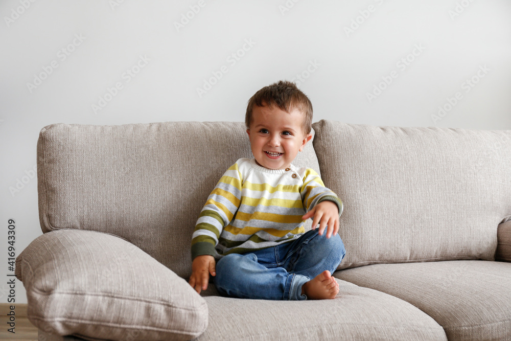 © Evrymmnt - Portrait of adorable little boy sitting on the textile couch and smiling. Happy toddler laughing and being playful at home. Barefoot kid in denim pants. Close up, copy space, background.