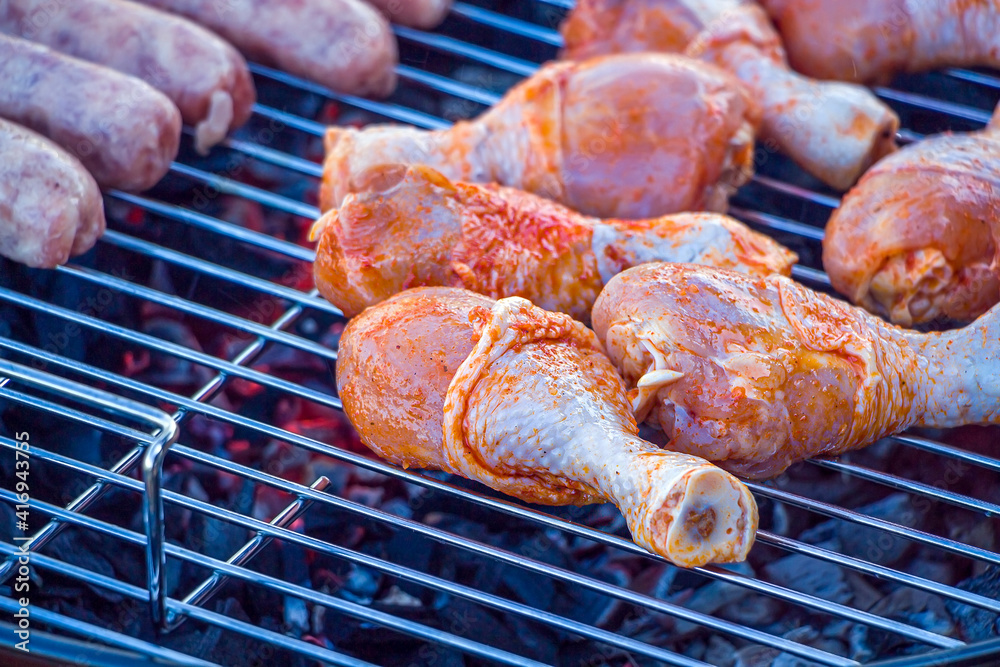 Process cooking chicken drumstick and sausages on barbecue grill outdoors. Picnic, eating outdoors. Metal barbecue grill (brazier). Raw meat.