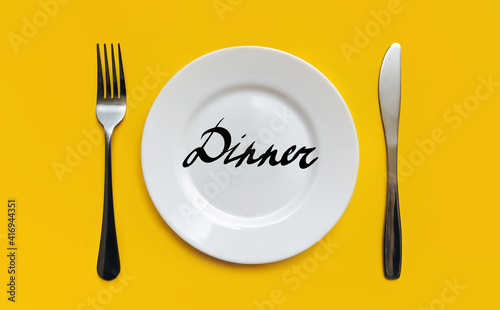 A white plate on which the word Dinner is written standing on a yellow tablecloth . The concept of a balanced diet, ration and medical fasting. Top view, white background, copy space.