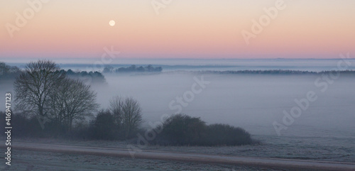 an early morning mist filled valley with the snow moon low in the sunrise sky 