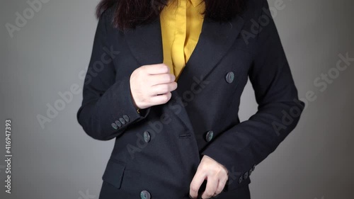 Front view on body part of young female in yellow blouse and black jacket. Close up of unrecognizable woman unbutton her jacket on grey background.
