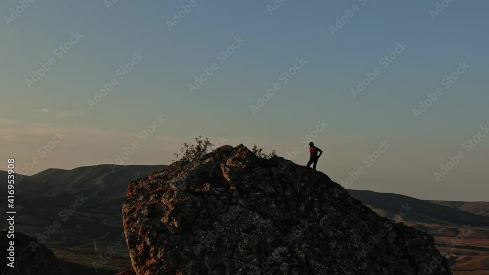 A Young Man in a Baseball Cap Climbs the Top of the Mountain Stops and Makes Victory Hand Gestures Against the Background of a Mountain Landscape with a Blue Sky and Sunset. Aerial view. Surveying by A Young Man in a Baseball Cap Climbs the Top of the Mountain Stops and Makes Victory Hand Gestures Against the Background of a Mountain Landscape with a Blue Sky and Sunset. Aerial view. Surveying by